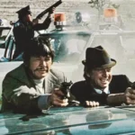 Ein Mann geht über Leichen - Filmkritik: -Titelbild: Männer zielen mit Pistolen hinter Auto, Polizeiwagen im Hintergrund.