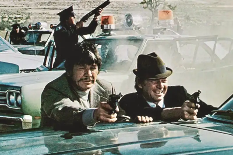 Ein Mann geht über Leichen - Filmkritik: -Titelbild: Männer zielen mit Pistolen hinter Auto, Polizeiwagen im Hintergrund.