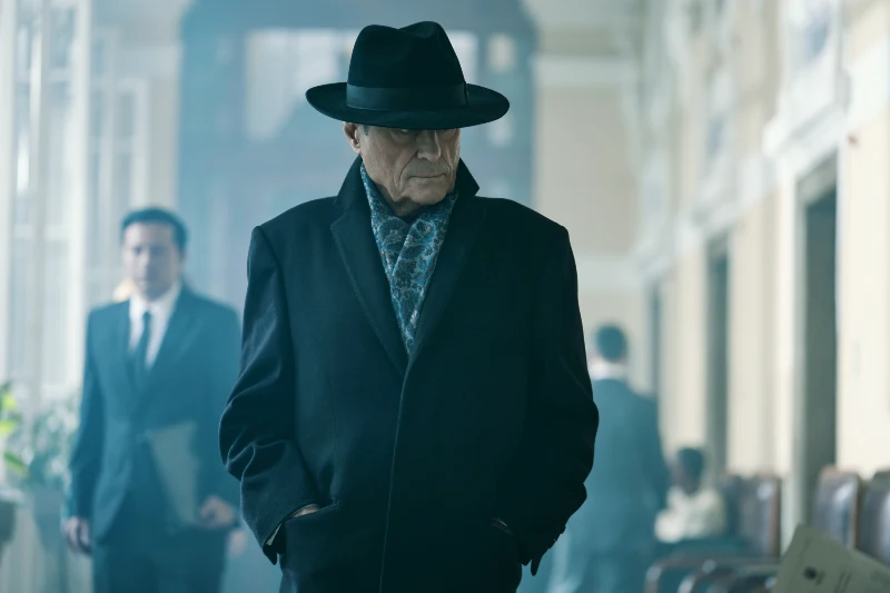 Darsteller Ian McShane mit Mantel und Hut in einem Gang mit Stühlen