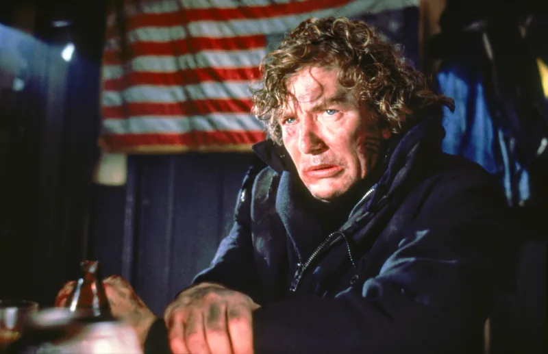 Ein erschöpfter Mann mit dreckigen Gesicht und schwarzen Jacke sitzt vor der US-Flagge - Filmszene Wolfen