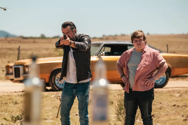 Darsteller Josh Duhammel und Jeremy Ray Taylor stehen vor einem Auto, während Duhammel mit einer Pistole auf Flaschen zielt - Filmszene London Calling