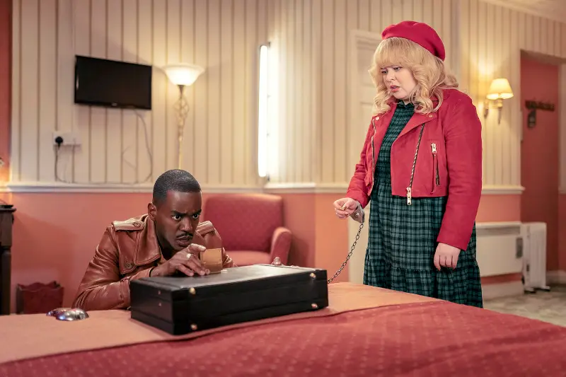 Ein Mann kniet vor einem Koffer, den er zu öffnen versucht, neben ihm eine Frau die mit Handschnellen an diesen Koffer gekettet ist - Szene aus Doctor Who Joy of the World