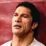 Dwayne Johnson geschminkt mit Make-Up Prothesen als MMA Kämpfer Mark Kerr, angelehnt an einer roten Wand