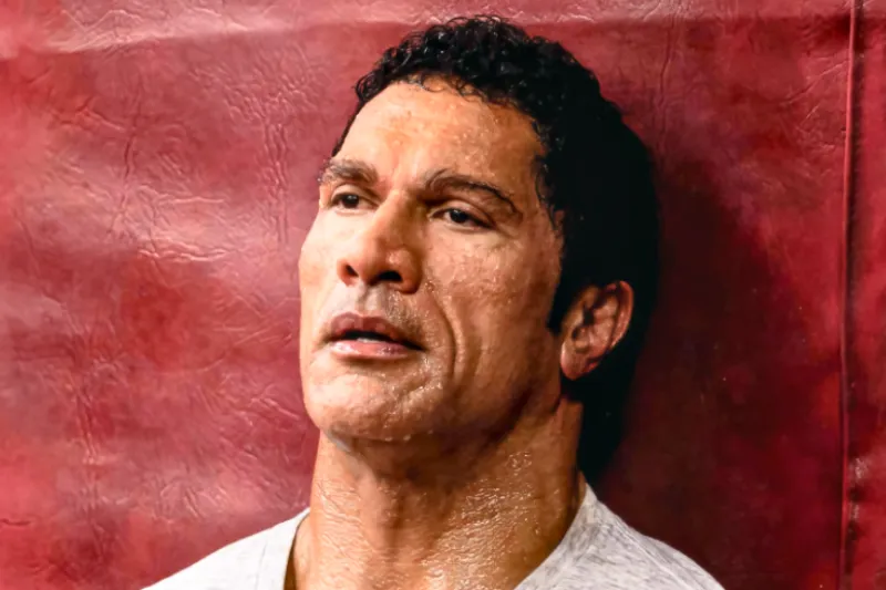 Dwayne Johnson geschminkt mit Make-Up Prothesen als MMA Kämpfer Mark Kerr, angelehnt an einer roten Wand