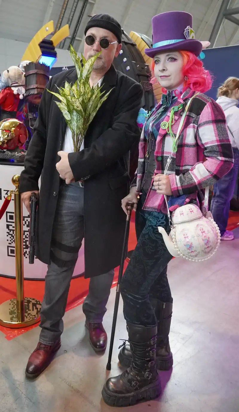 Ein Mann mit Sonnenbrille, langen Mantel, Pistole und Blumentopf in der Hand, daneben der verrückte Hutmacher aus Alice im Wunderland