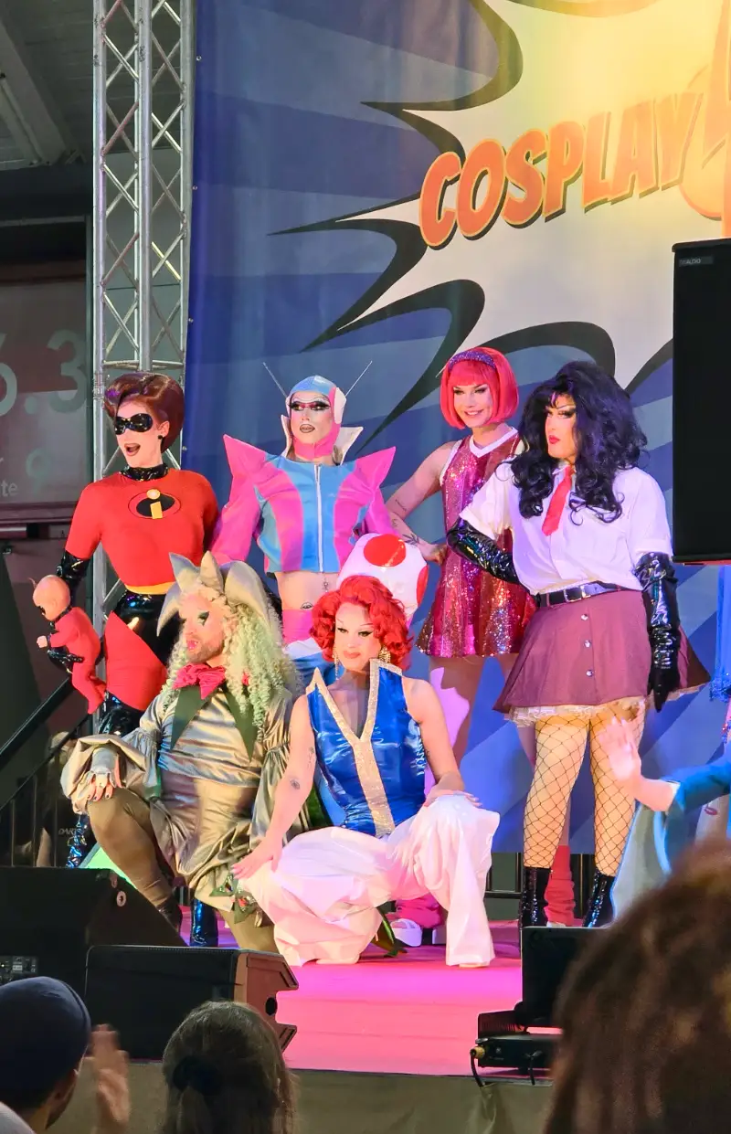 Eine Gruppe bunt gekleideter Drag Queens auf einer Bühne in einer Messehalle