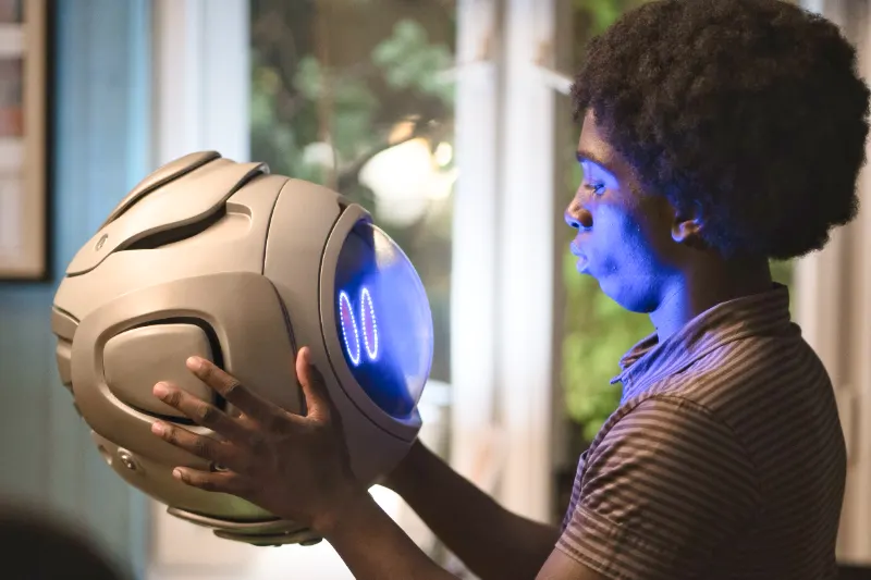 Ein Junge hält einen runden Roboter mit Gesicht in den Händen - Szene aus Momo 2025 araloyin oshunremi als gino mit bb bot