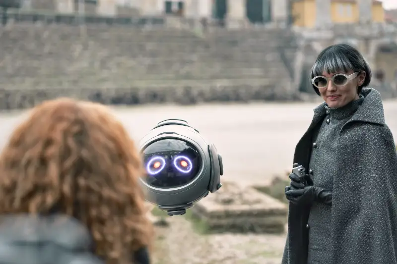 Frau spricht mit schwebendem Roboter im Freien vor rothaarigen Mädchen - Szene Momo 2025