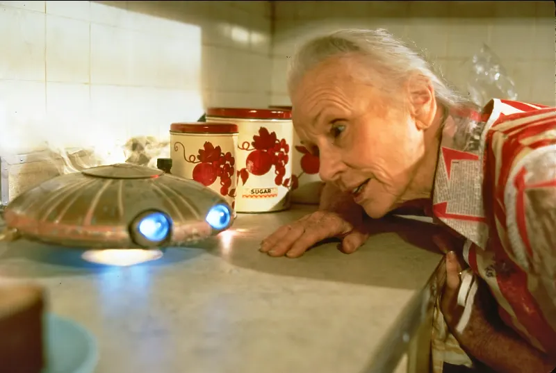 Eine Frau (Darstellerin Jessica Tandy) beugt sich auf ihrem Küchentresen zu einem kleine UFO hinunter - Filmszene