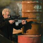 Mel Gibson als Bowdrie mit Sturmgewehr hinter Ölfässern in Hunting Season