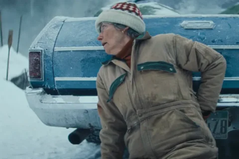 Frau in Winterjacke blickt alarmiert hinter blaues Auto im Schnee.