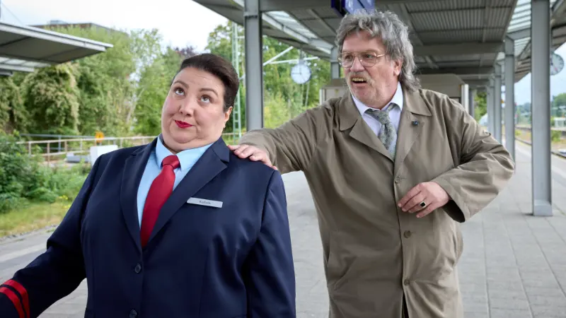 Mann (Hape Kerkeling) im Trenchcoat spricht mit genervter Bahnmitarbeiterin am Bahnsteig.