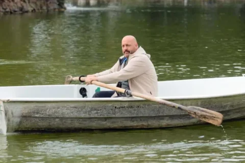 Kahlköpfiger Mann (Kevin James) rudert ein Boot auf ruhigem See