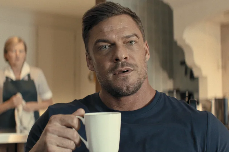Mann hält eine weiße Tasse in der Hand; im Hintergrund steht eine Person in einer Küche