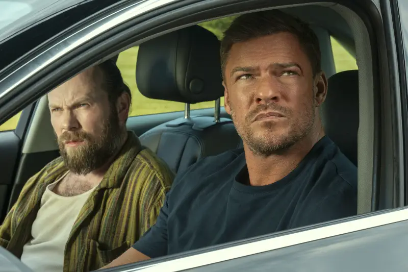 Alan Ritchson und ein Mann mit Vollbart blicken aufmerksam aus dem Fenster eines Autos
