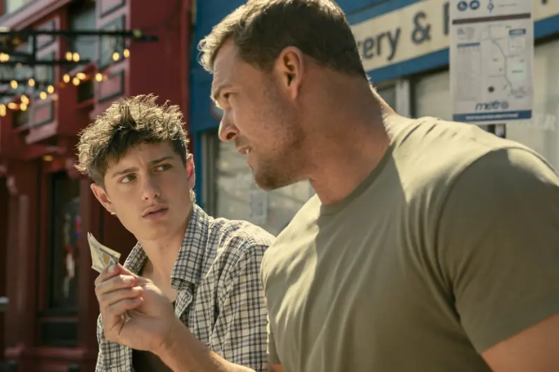 Alan Ritchsons als Reacher spricht intensiv mit einem jüngeren Mann und hält einen Geldschein in der Hand.